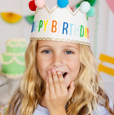 Child wearing a colorful 'Happy Birthday' crown with a blurred birthday cake in the background.