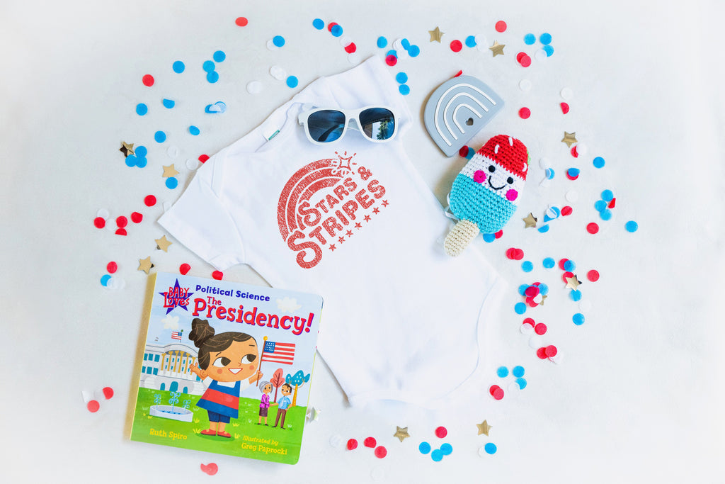 Little Holiday The Babe Box for the 4th of July. Includes Baby Loves, Political Science, The Presidency baby board book, white Babiator sunglasses, Pebble Child red, white and blue popsicle rattle, Getting Sew Crafty silicone rainbow teether, and a custom stars and stripes onesie design. 4th of July confetti.